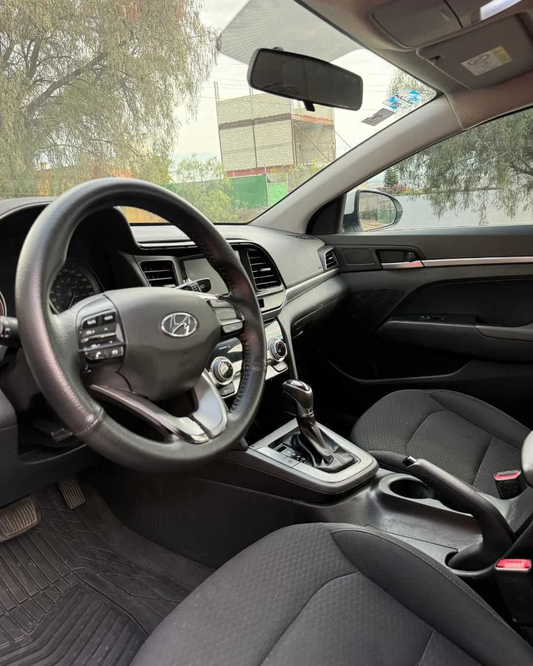 Interior de un coche gris, vista del volante, tablero, consola central y asientos, con un paisaje exterior visible por las ventanas.