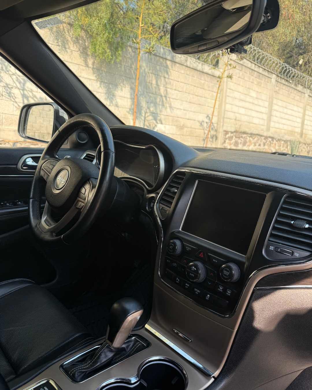 Interior de un automóvil tipo SUV, vista del volante, tablero, pantalla central y consola con palanca de cambios, con fondo de muro y árboles vistos a través del parabrisas.