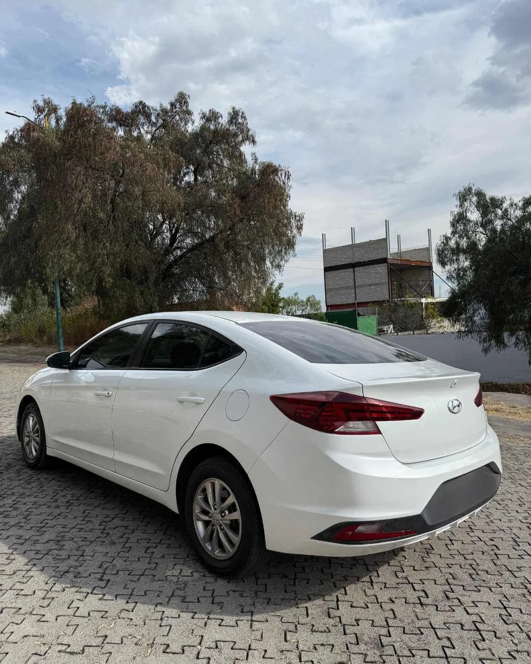 Auto sedán blanco estacionado sobre pavimento de bloques en área al aire libre, con árboles y estructura en construcción al fondo y cielo nublado.