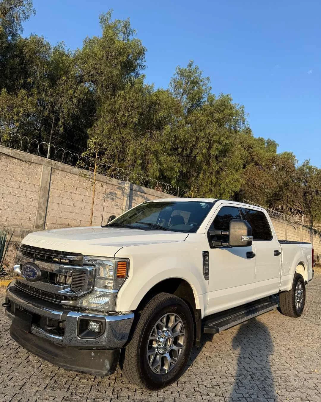 Una camioneta Ford blanca estacionada en una calle adoquinada, con una cerca de alambre de púas y árboles verdes en el fondo bajo un cielo azul despejado.