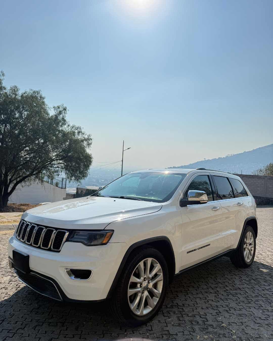 Coche SUV color blanco estacionado en una zona con pavimento de piedra, con colina y árboles al fondo, cielo despejado y sol brillante.