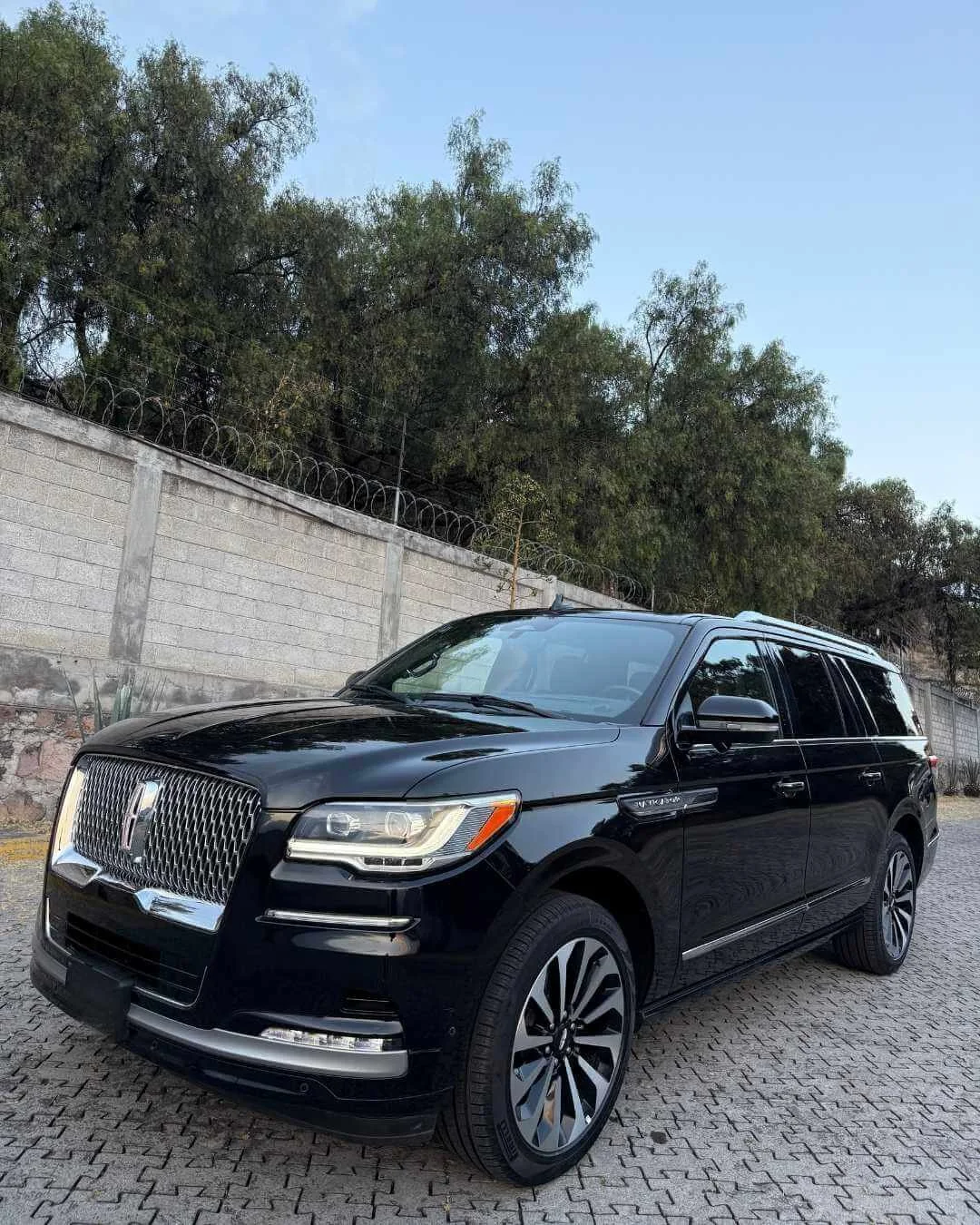 Un coche negro tipo SUV estacionado sobre un pavimento de piedra, con un muro y árboles verdes en el fondo bajo un cielo despejado.