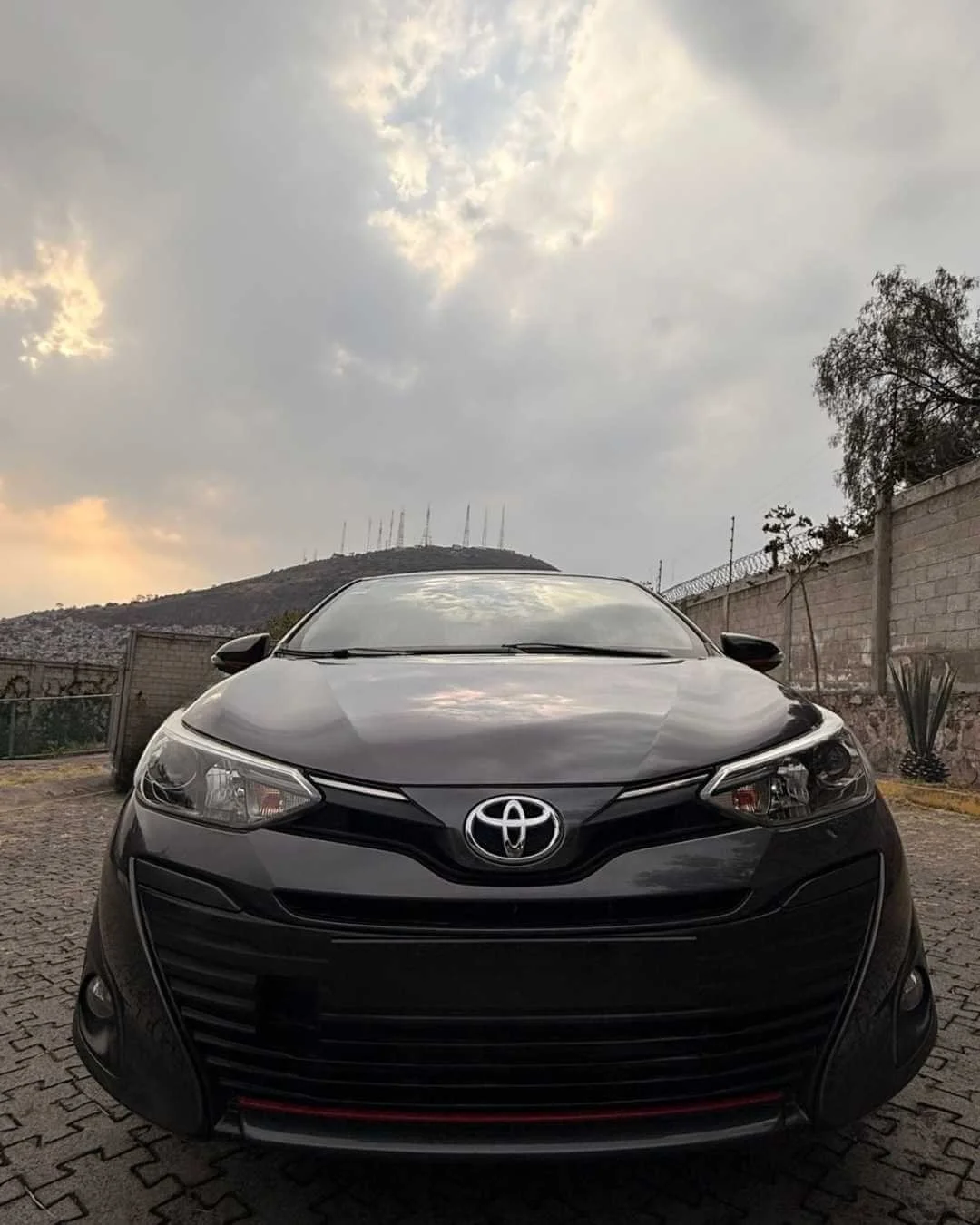 Auto negro estacionado en una superficie de piedra, con fondo de colina y cielo nublado.