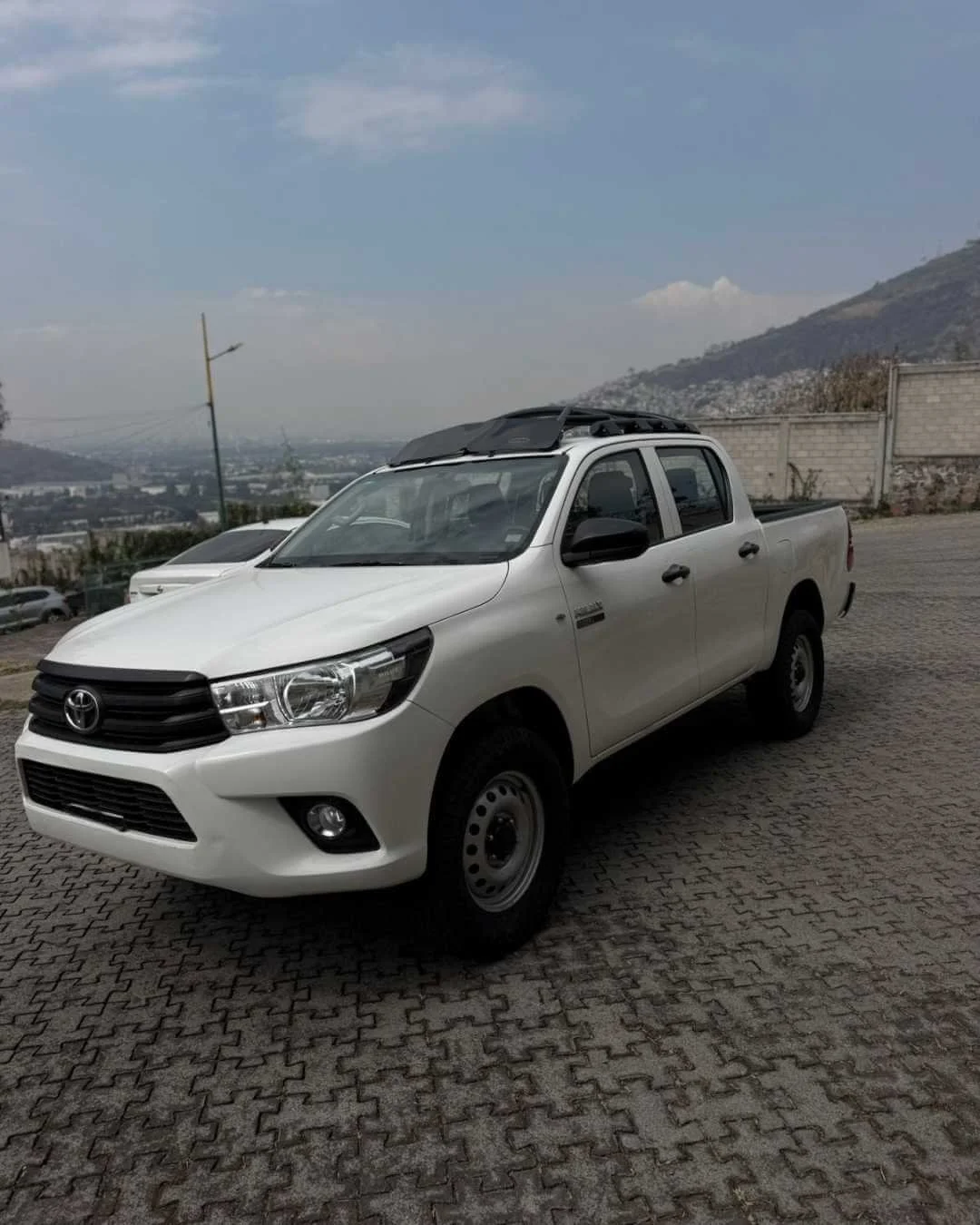 Camión Toyota blanco estacionado en un área pavimentada con montañas y cielo nublado de fondo.