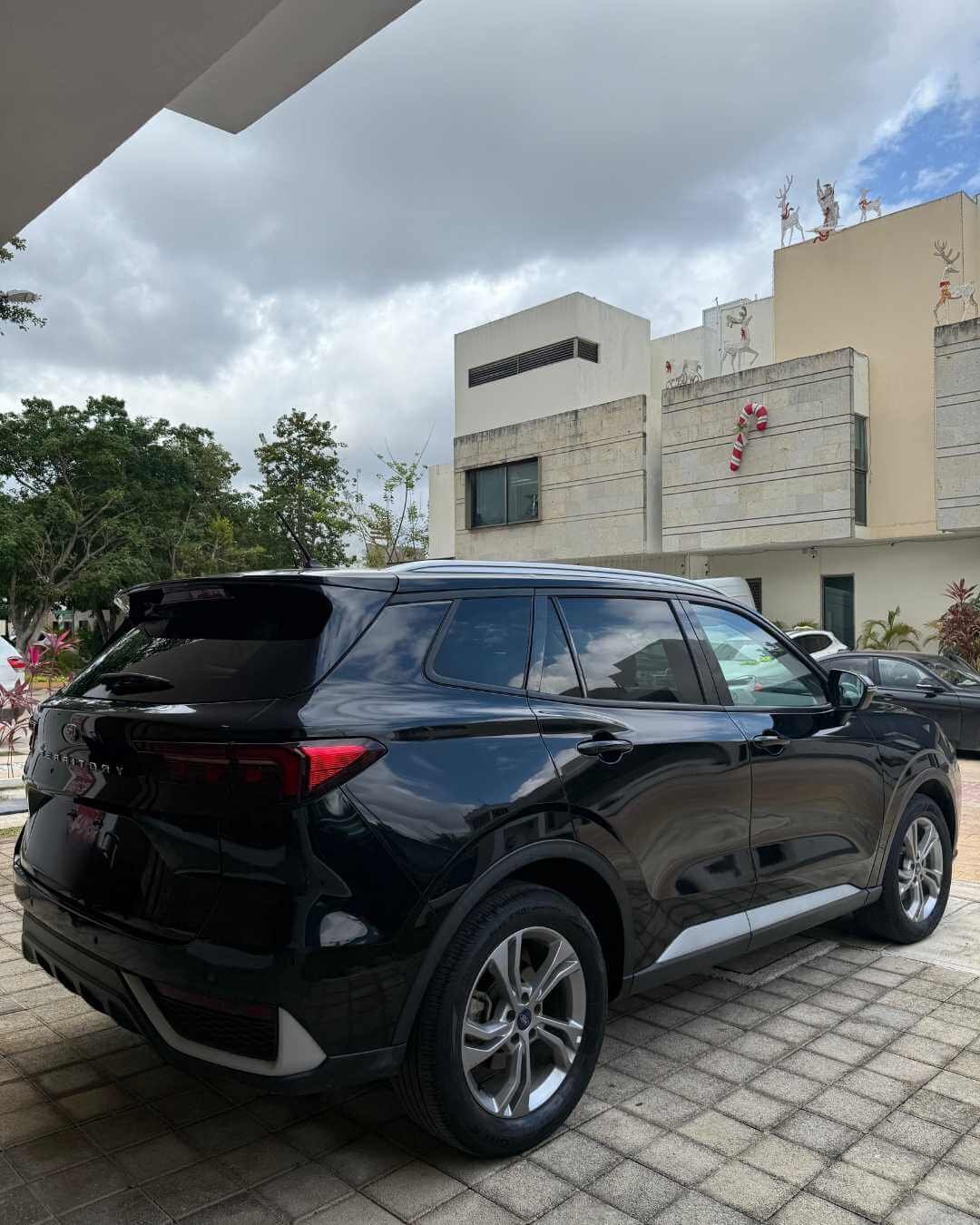 Cochera con un automóvil SUV negro y un edificio moderno decorado con motivos navideños, incluyendo un adorno en forma de bastón de caramelo y renos en la azotea. Árboles y otros vehículos también visibles.