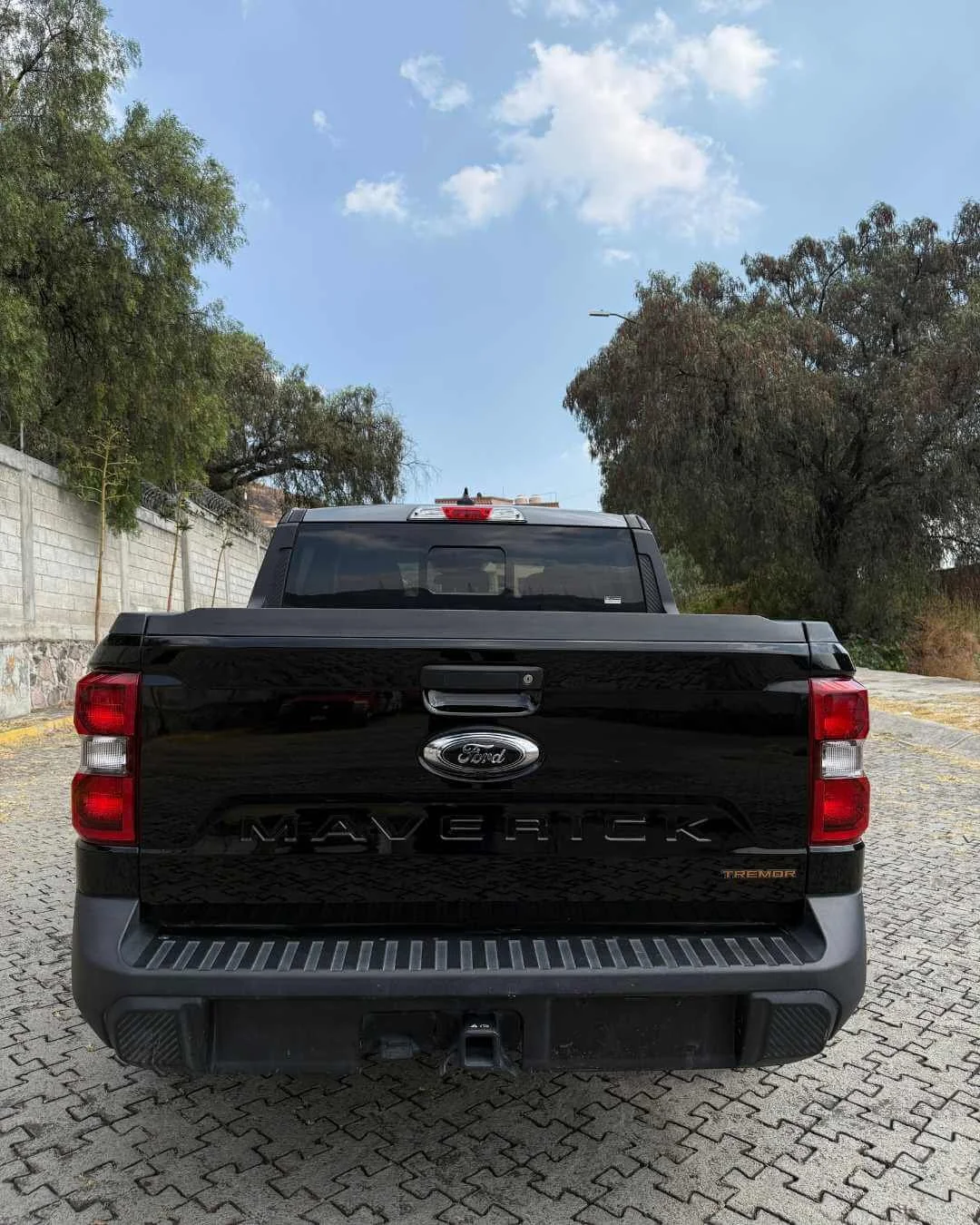 Camioneta negra Ford Maverick TREMOR estacionada sobre una calle de piedra, con árboles y muro de fondo y cielo despejado con algunas nubes.