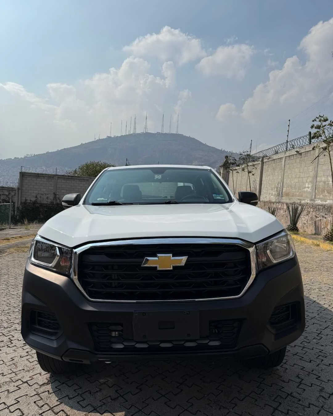 Frontal de un auto Chevrolet blanco estacionado en un área de pavimento de piedra con un muro de fondo y una colina con antenas en el horizonte, bajo cielo con nubes.