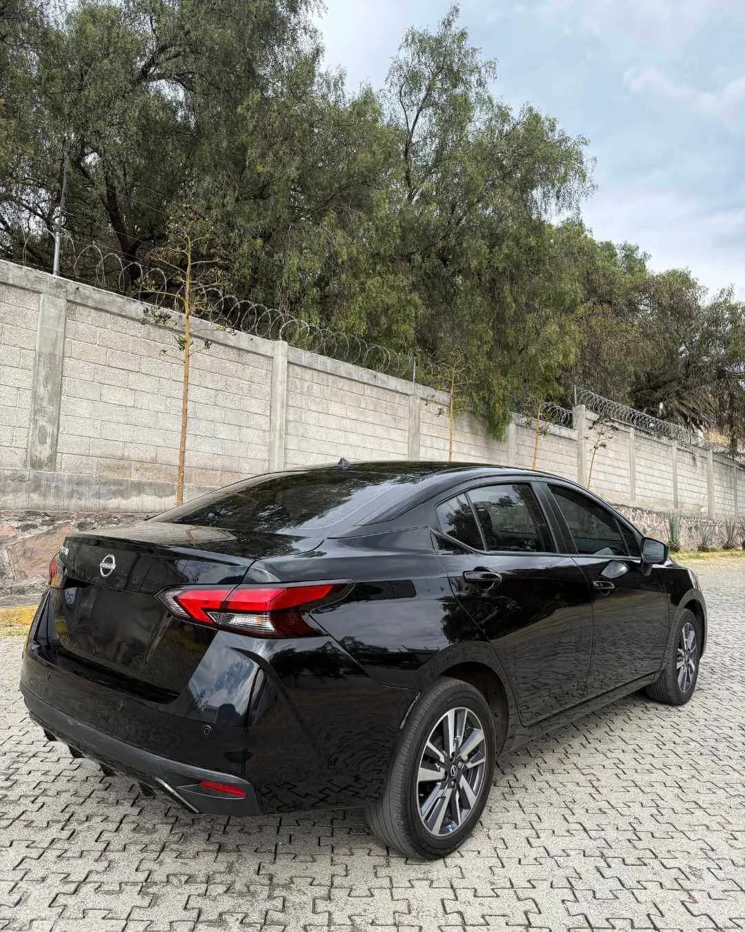Coche negro estacionado en un área pavimentada con un muro y árboles en el fondo.