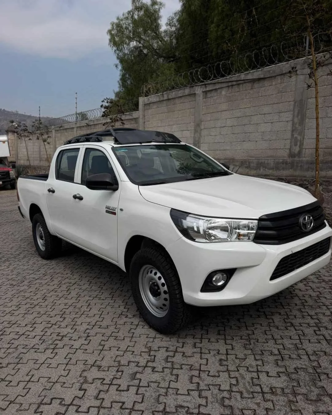 Camioneta blanca Toyota en estacionamiento con muro de concreto y alambre de púas en el fondo.