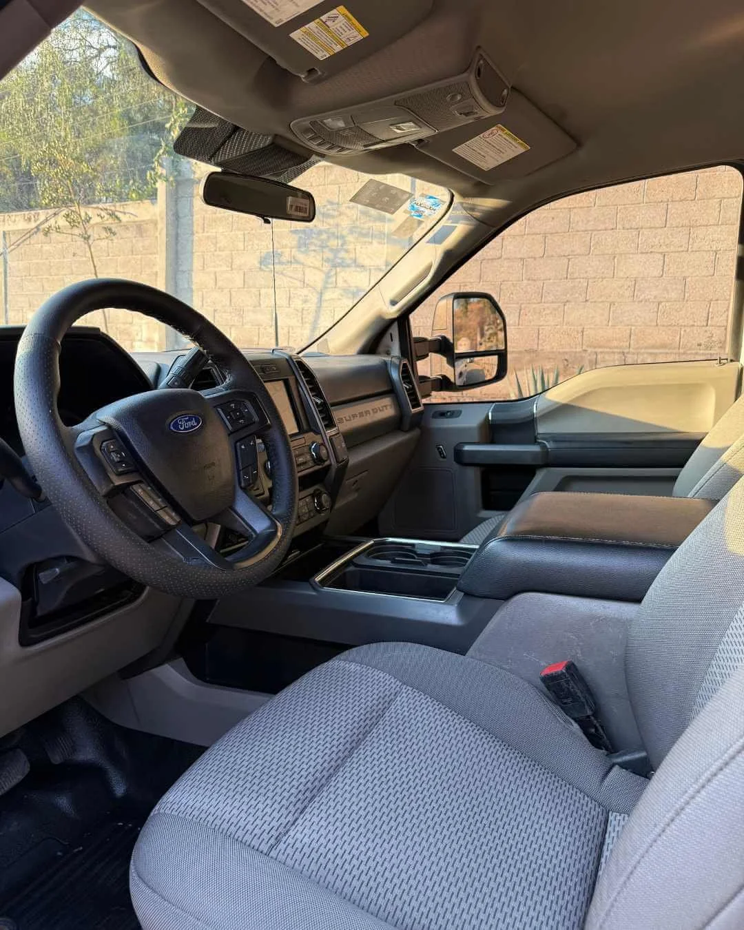 Interior de una camioneta Ford con asiento del conductor y tablero, en un entorno con pared de ladrillos exteriores y árboles visibles a través del parabrisas.