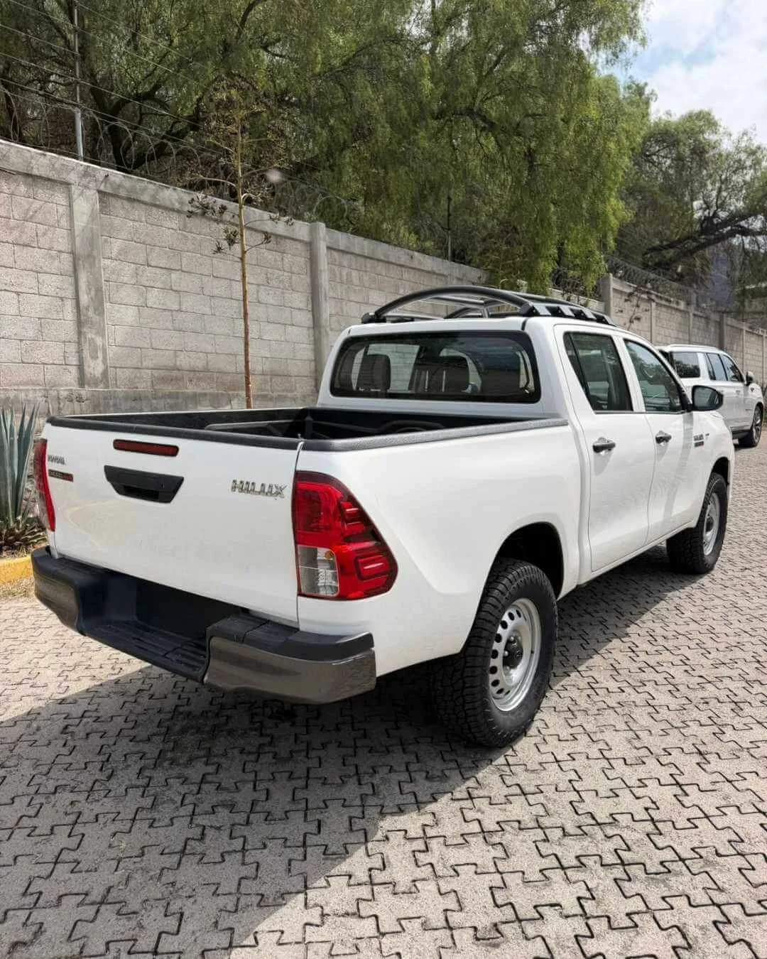 Camioneta pickup blanca estacionada en un estacionamiento con muro de cemento y árboles en el fondo.