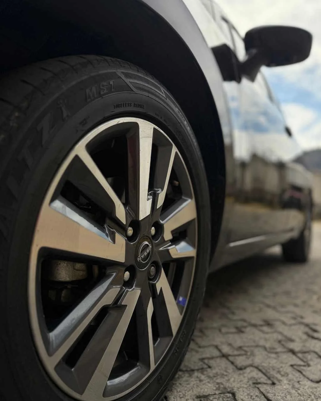 Detalle de una rueda de carro con llanta estilo deportivo en un coche negro, estacionado sobre pavimento de piedra.