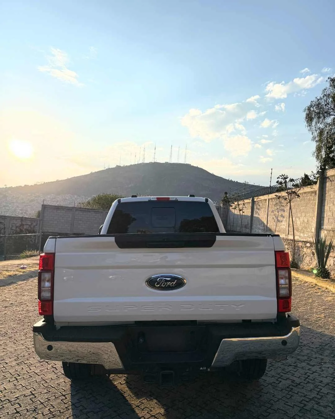 Camioneta Ford de color blanca, vista desde atrás, estacionada en un camino con muro de ladrillos a la derecha y un cerro con antenas en el fondo, al atardecer.