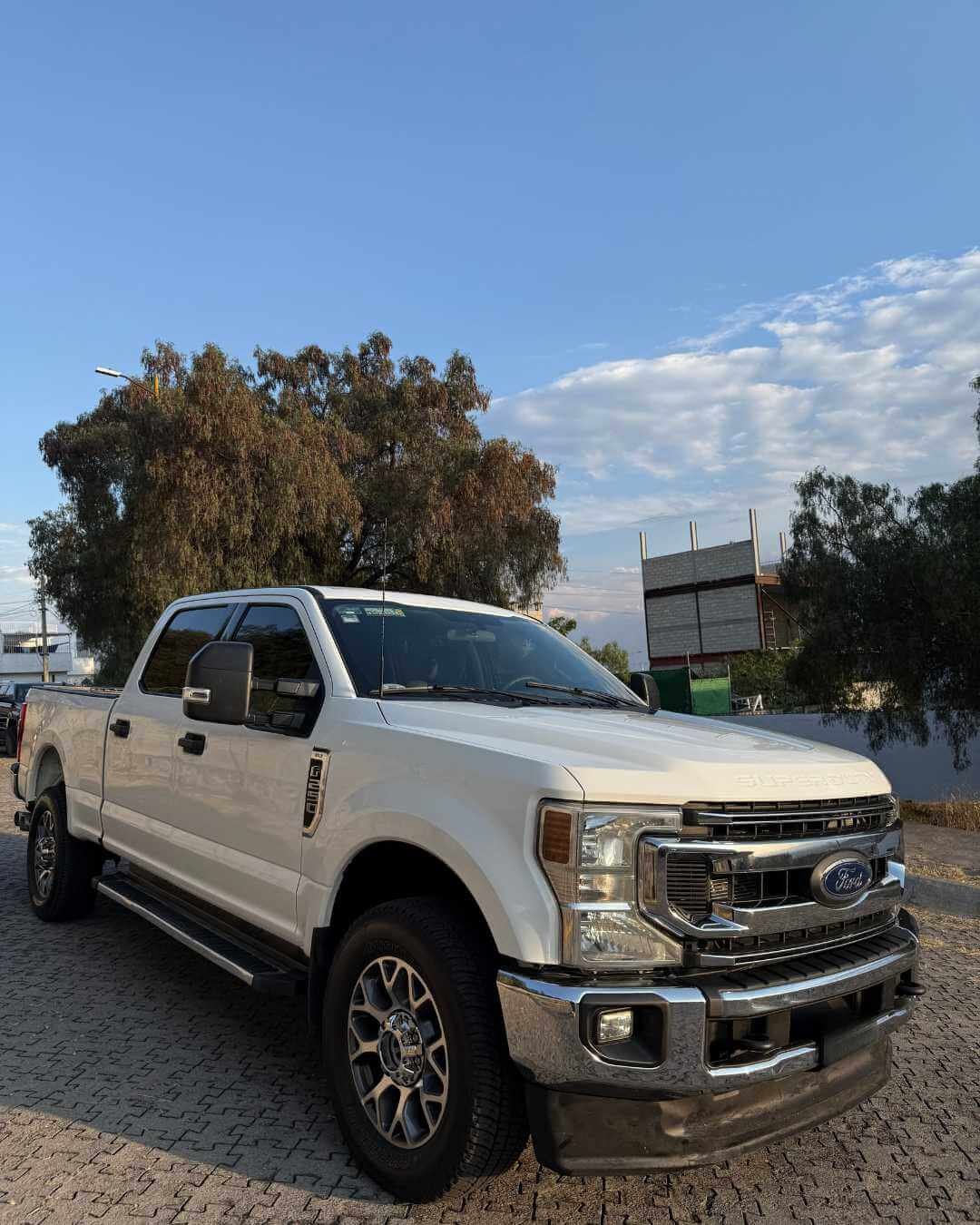 Camioneta Ford blanca estacionada sobre pavimento de ladrillos en un entorno al aire libre con árboles y cielo con nubes en el fondo.