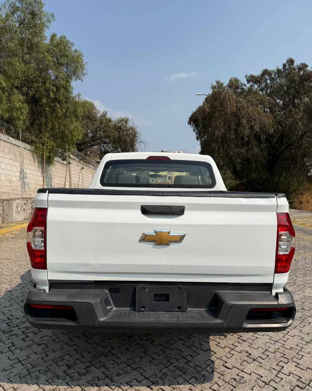 Imagen de la parte trasera de una camioneta blanca con un logotipo dorado de Chevrolet, estacionada en una calle con árboles a ambos lados y un muro de bloques de concreto a la izquierda.