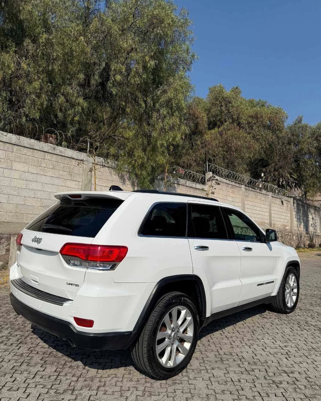 Un SUV blanco estacionado en un área pavimentada con árboles y una pared de fondo.