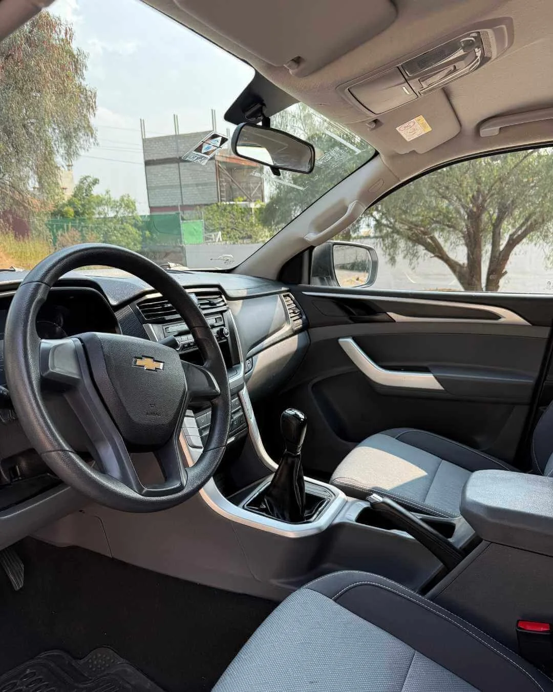 Interior de un coche con volante, palanca de cambios y asiento. Se ven ventanas con vista a árboles y el exterior.