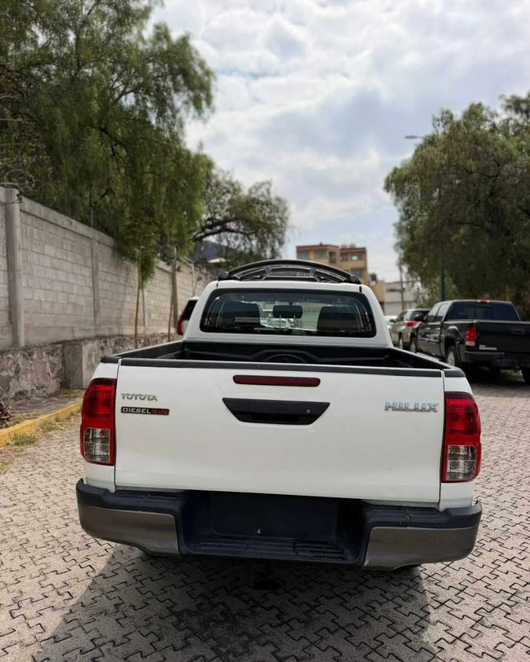 Camioneta Toyota Hilux blanca vista desde atrás en un estacionamiento, con árboles y edificios al fondo y cielo nublado.