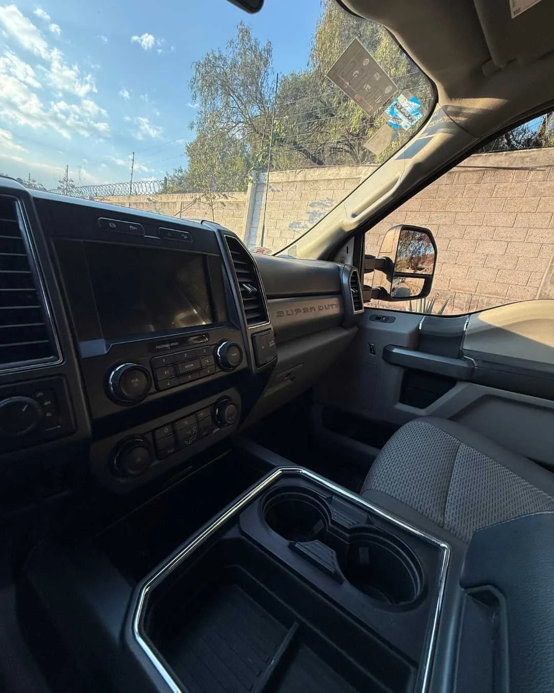 Vista del interior de una camioneta negra con tablero de instrumentos y consola central con compartimientos y porta vasos, ventanillas y un espejo lateral, con un muro y árboles en el fondo.