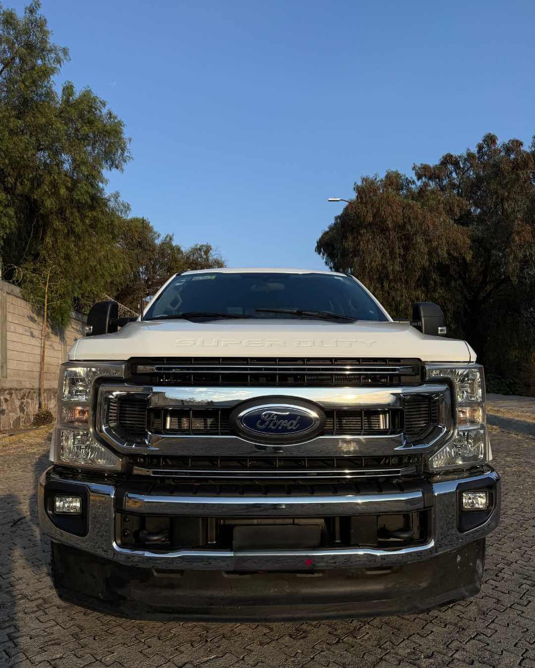 Camioneta Ford Super Duty blanca frente vista, en un camino de piedra, con árboles y cielo despejado en el fondo.
