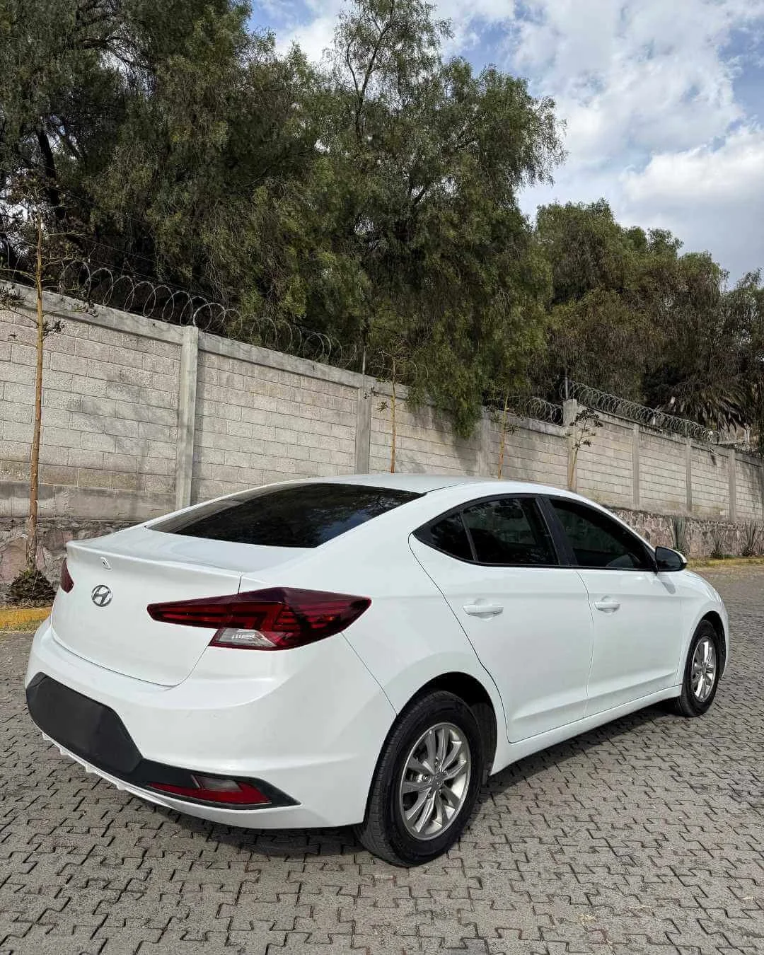 Automóvil sedán blanco estacionado en un área pavimentada, con muro de bloques y cercas de espiral en el fondo, y árboles al fondo bajo un cielo parcialmente nublado.
