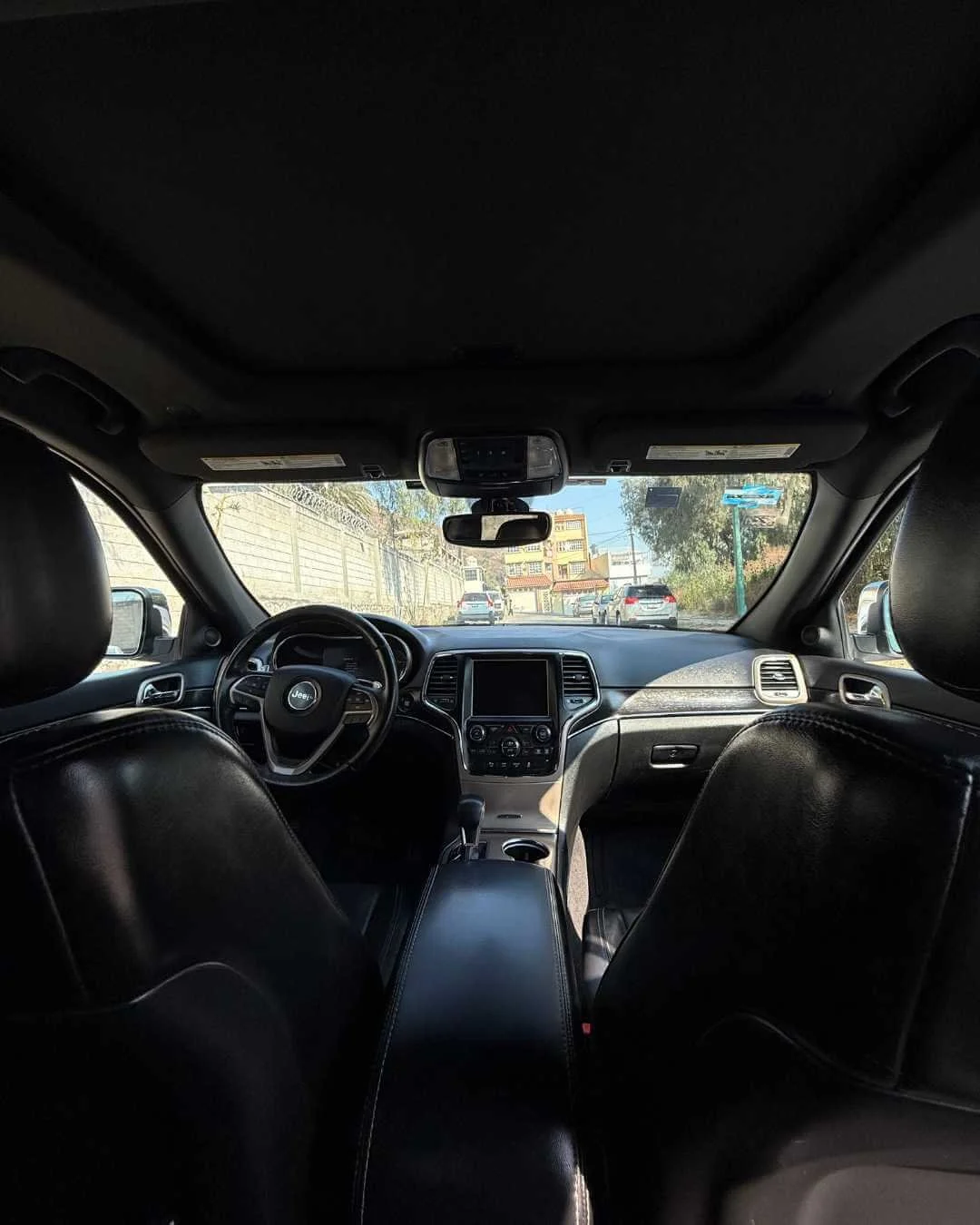 Interior de un auto con asientos de cuero negro, tablero con pantalla y volante con logo Jeep, vista hacia la calle con autos y edificios a través del parabrisas.