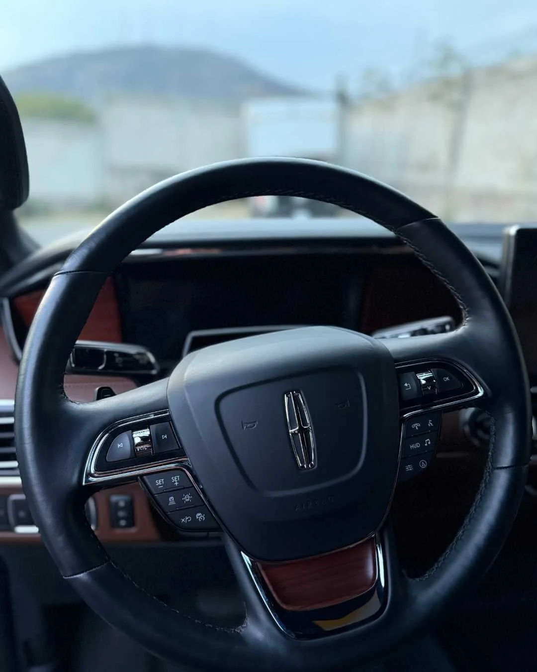 Volante de coche con logotipo de Lincoln en el centro, visible tablero y algunas advertencias en el fondo, con paisaje de fondo a través del parabrisas.