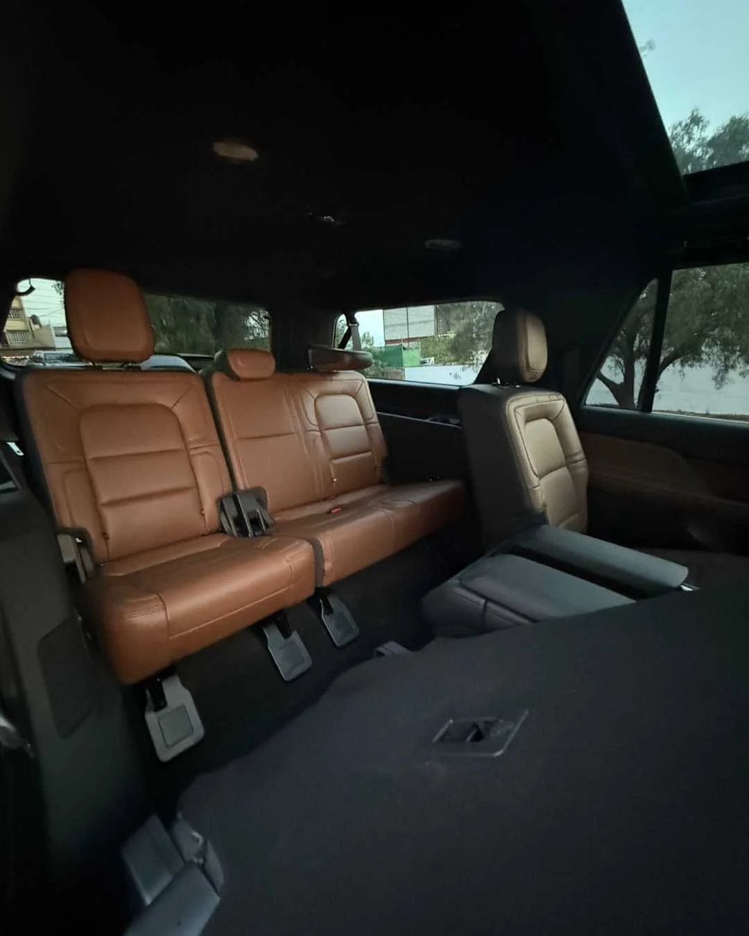 Interior de un vehículo con asientos de cuero marrón y negro, vista desde el maletero, con ventanas que muestran árboles y edificios en el fondo.