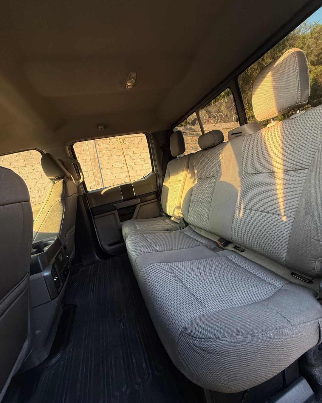 Interior de un vehículo con asientos de tela color gris claro y ventanas, con luz del sol entrando, y una pared de ladrillos visible desde el vehículo.