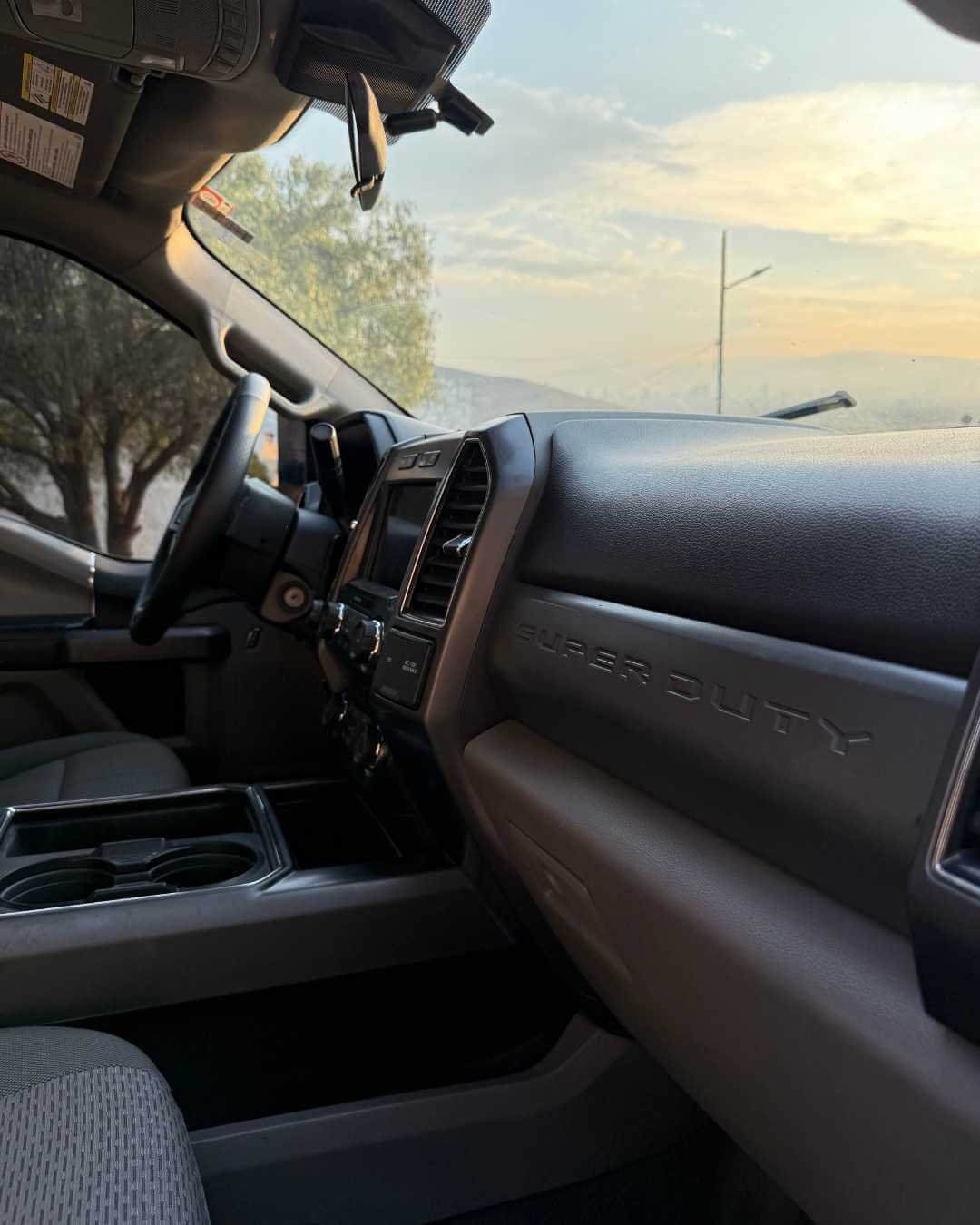 Interiores de una camioneta, vista desde el asiento del pasajero, mostrando el tablero y el volante, con el logo 'Elder Duty' y un paisaje con árboles, cielo y un poste de luz al fondo.