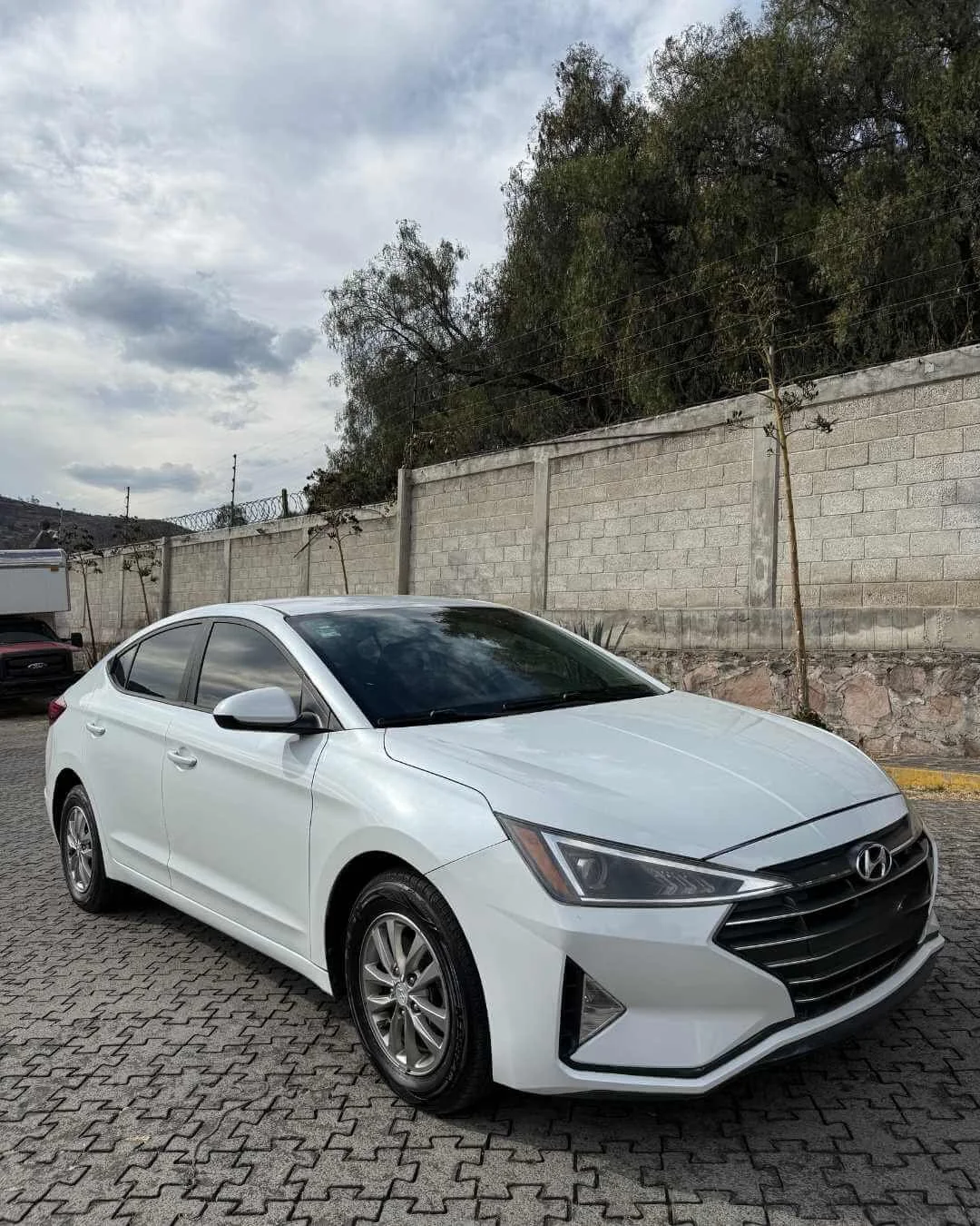 Coche sedán blanco marca Hyundai estacionado en un lugar al aire libre con árboles y muro de piedra en el fondo, cielo nublado.