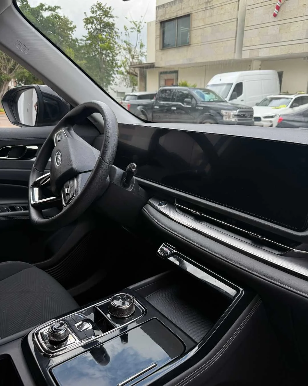 Vista del interior de un automóvil con volante y consola central, y vista exterior de otros autos estacionados y un edificio de fondo.