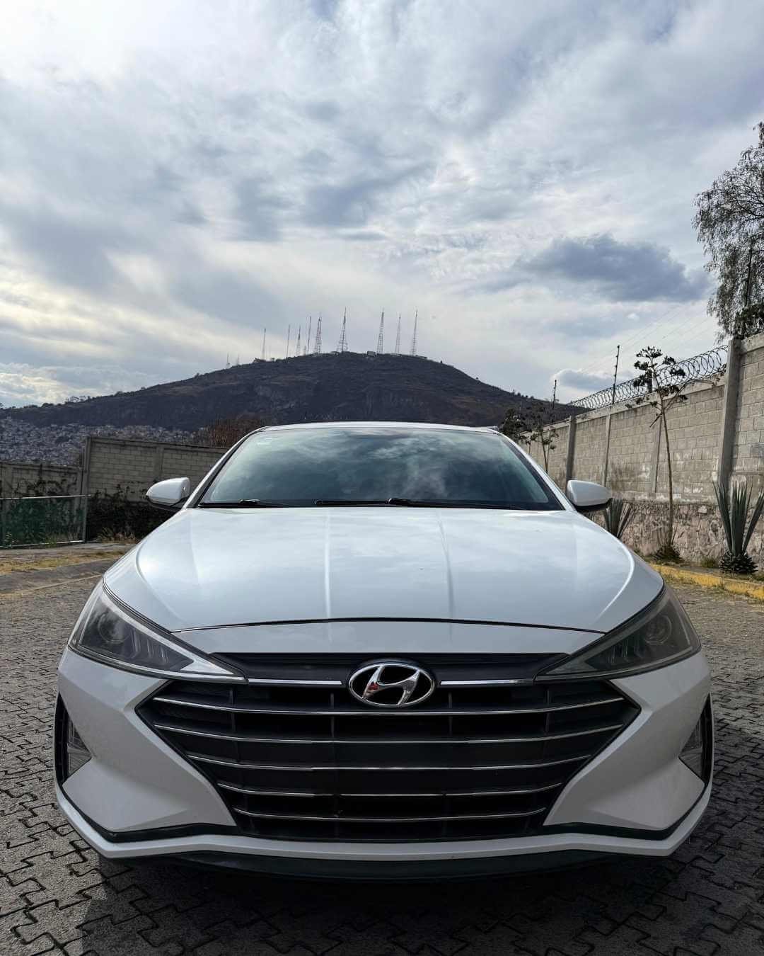 Coche blanco Hyundai estacionado en un parque con un cerro y antenas en el fondo, cielo nublado.