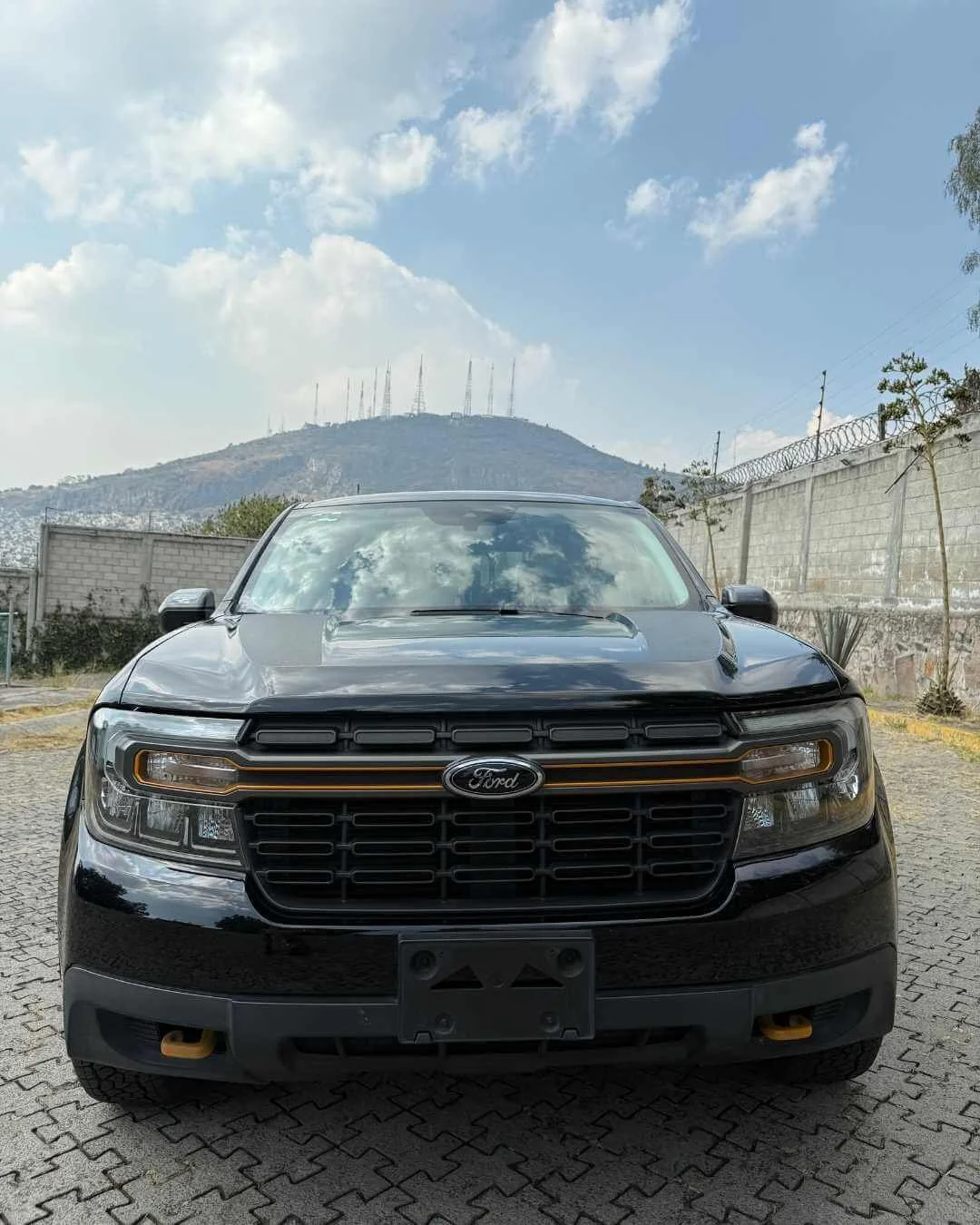 Auto negro estacionado sobre pavimento de piedra, con colinas y antenas en el fondo bajo cielo parcialmente nublado.