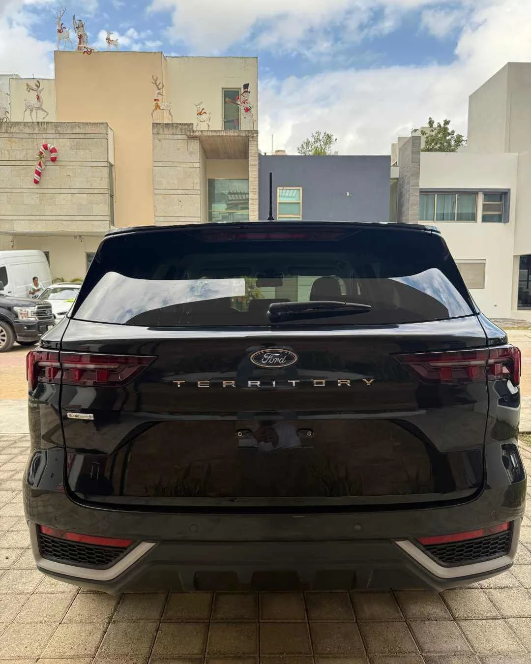 Automóvil Ford Territory negro estacionado en un área pavimentada, con edificios residenciales de fondo decorados con adornos navideños y figuras de renos en el techo.