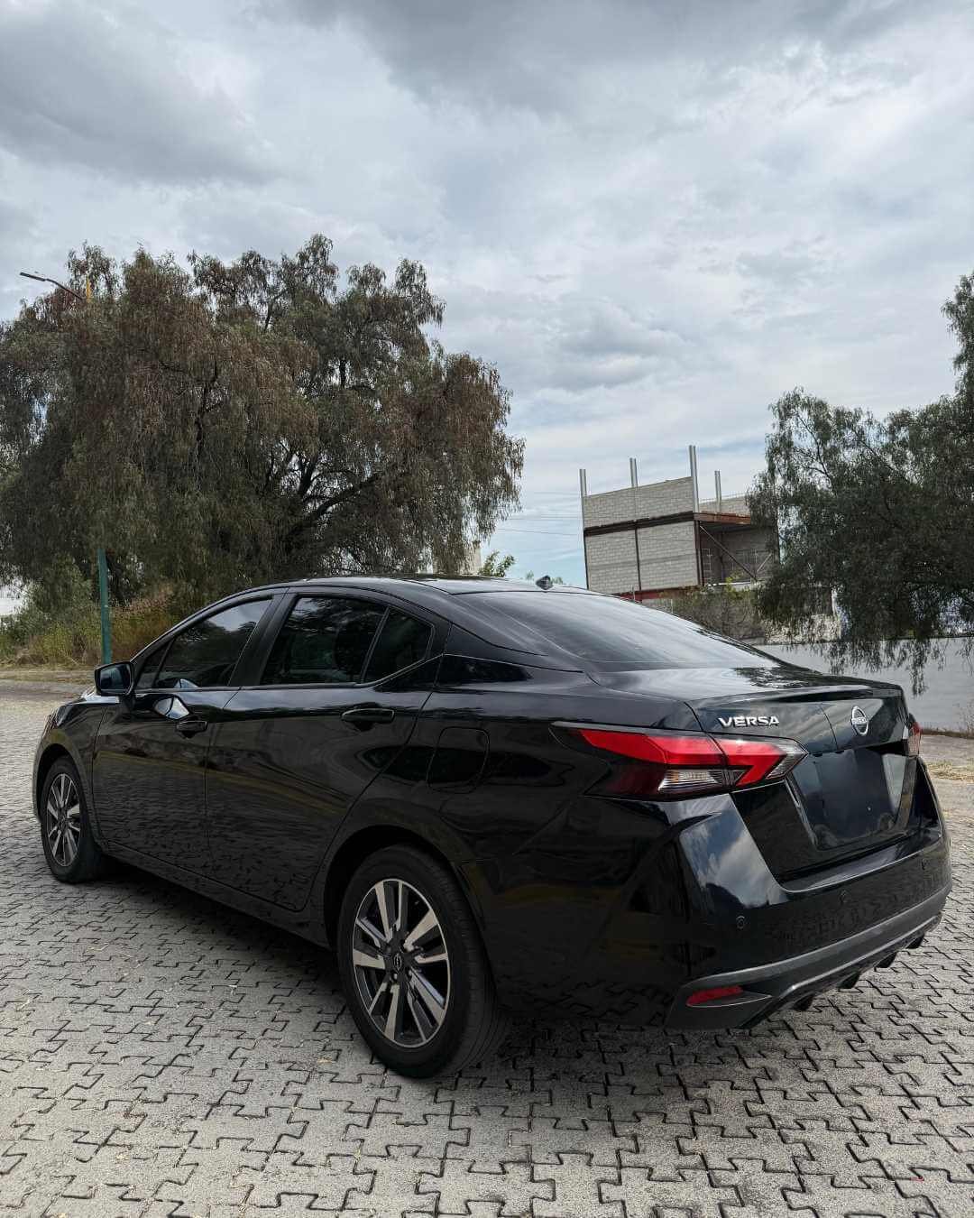 Auto negro Nissan Versa estacionado en un área pavimentada con árboles y cielo nublado en el fondo.