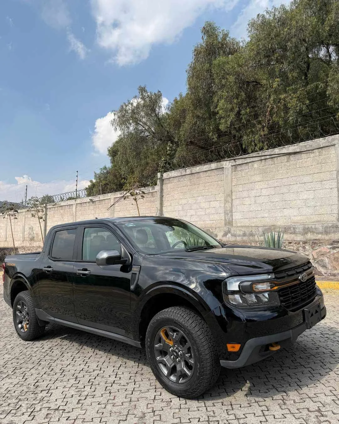 Coche negro tipo pickup estacionado en un área con pavimento de piedra, cerca de un muro de bloques con alambre de púas, árboles al fondo y cielo parcialmente nublado.