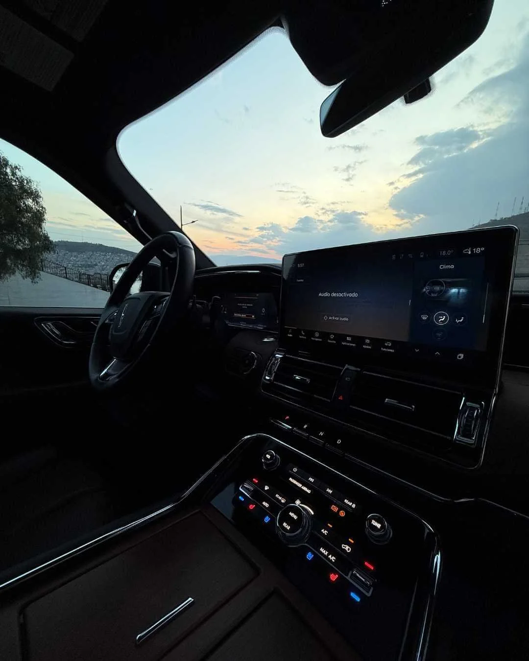Vista del interior de un coche en la noche, con un tablero digital y controles de clima iluminados, y una ventana que muestra un atardecer al aire libre.