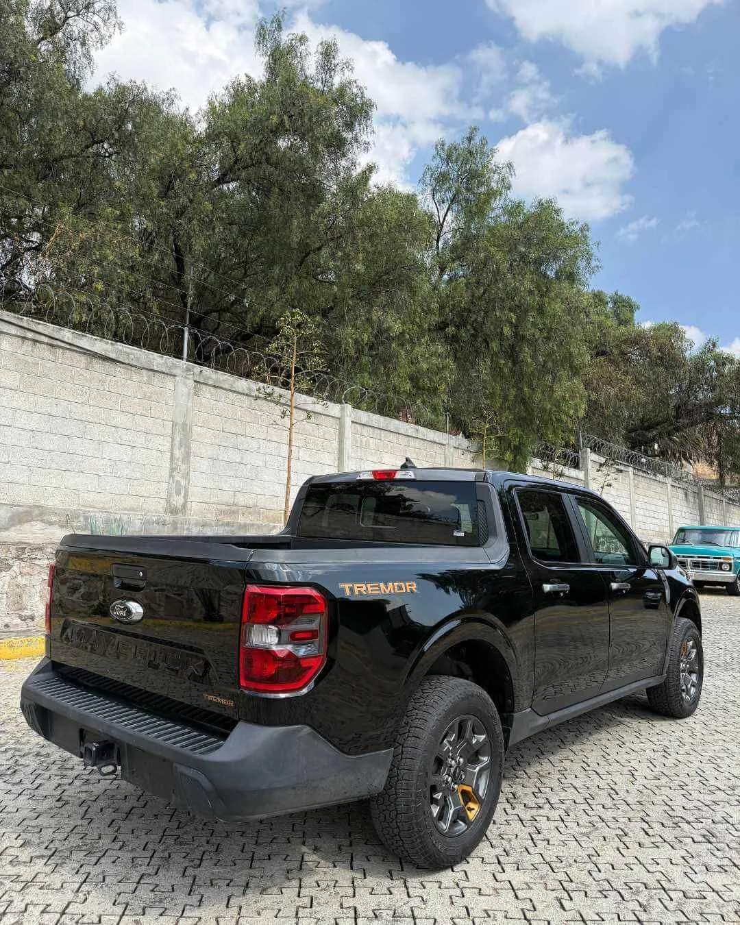 Camioneta negra en un estacionamiento, con letras doradas que dicen 'TREMOR', y un fondo de árboles y cielo con nubes.