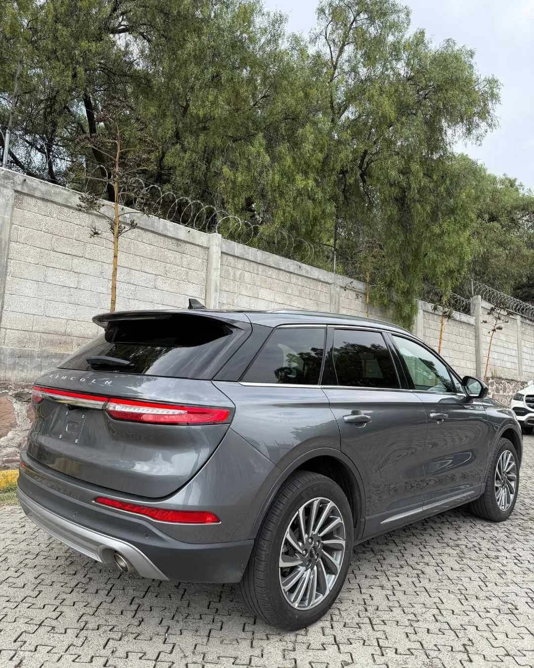 Auto SUV gris estacionado sobre pavimento de piedra, cerca de una pared con concertina, árboles verdes al fondo y cielo nublado.