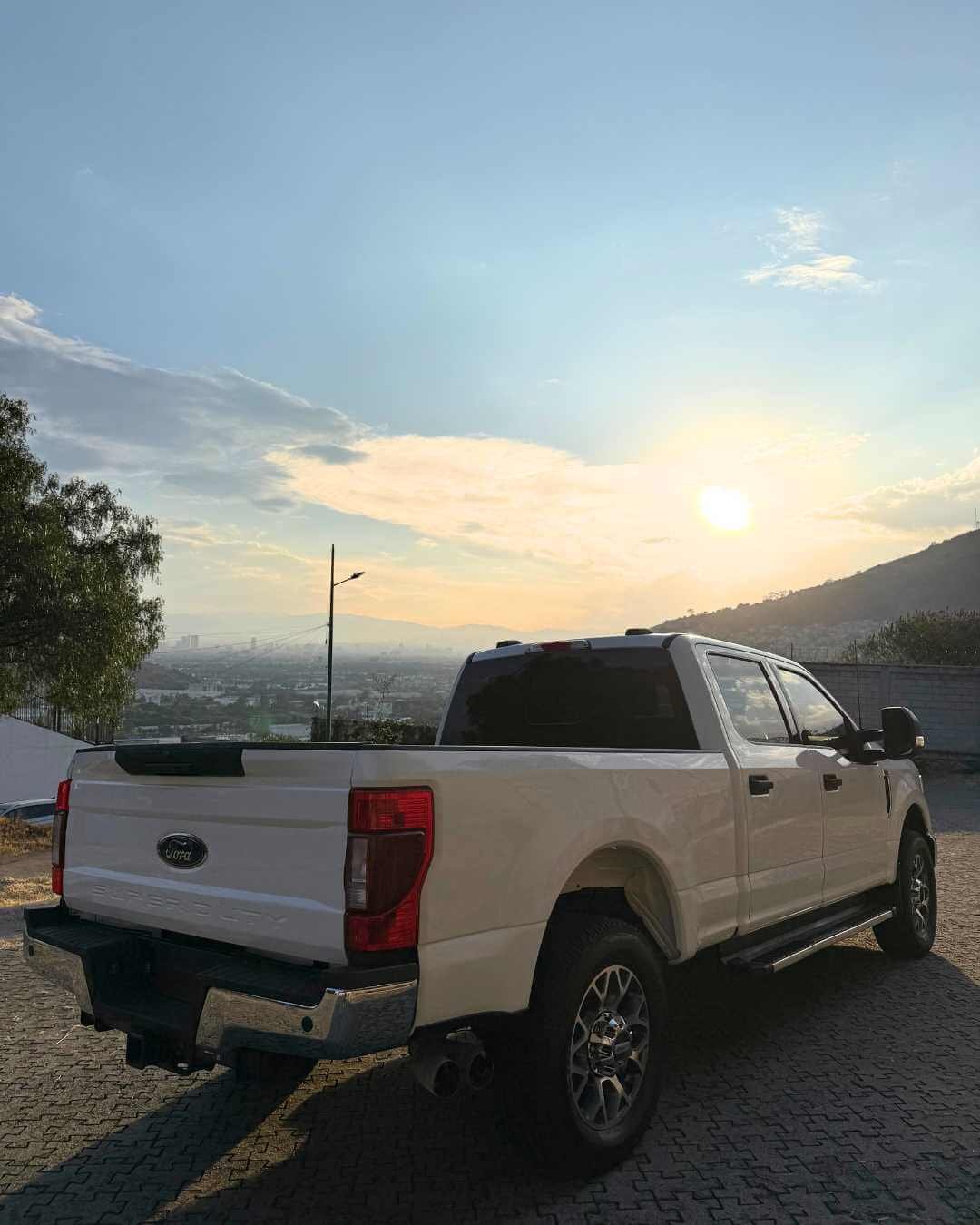 Camioneta blanca en un estacionamiento al amanecer o atardecer, con fondo de colinas y cielo parcialmente nublado.