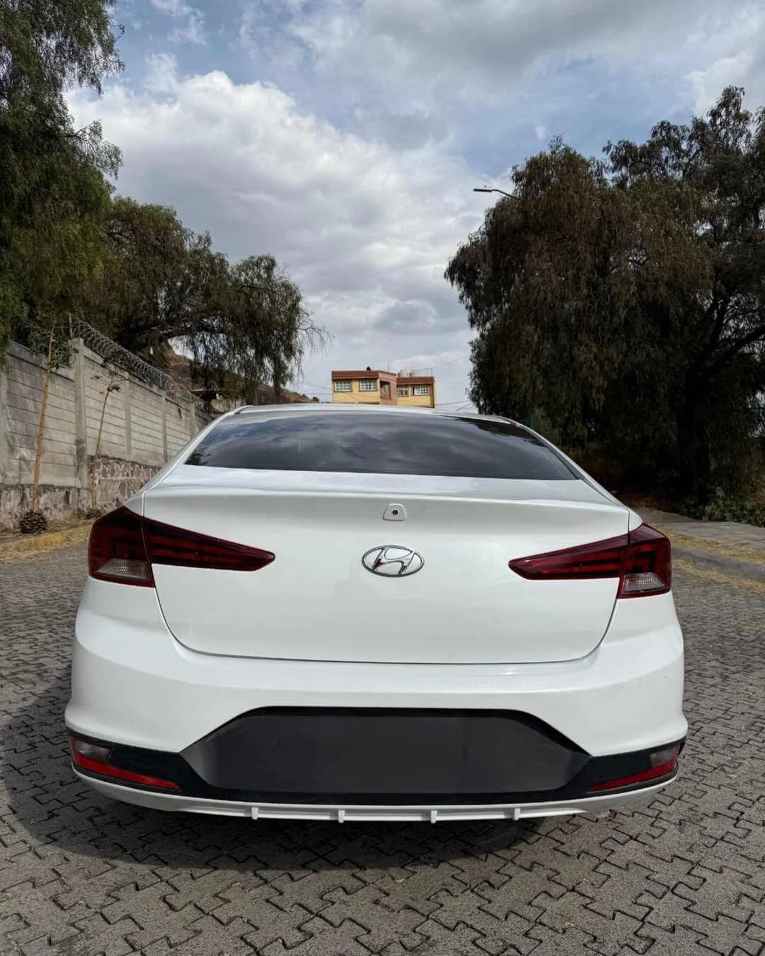 Auto blanco de perspectiva trasera en una calle de pavimento de bloques, con árboles y un muro a la izquierda y un edificio en el fondo, cielo nublado.