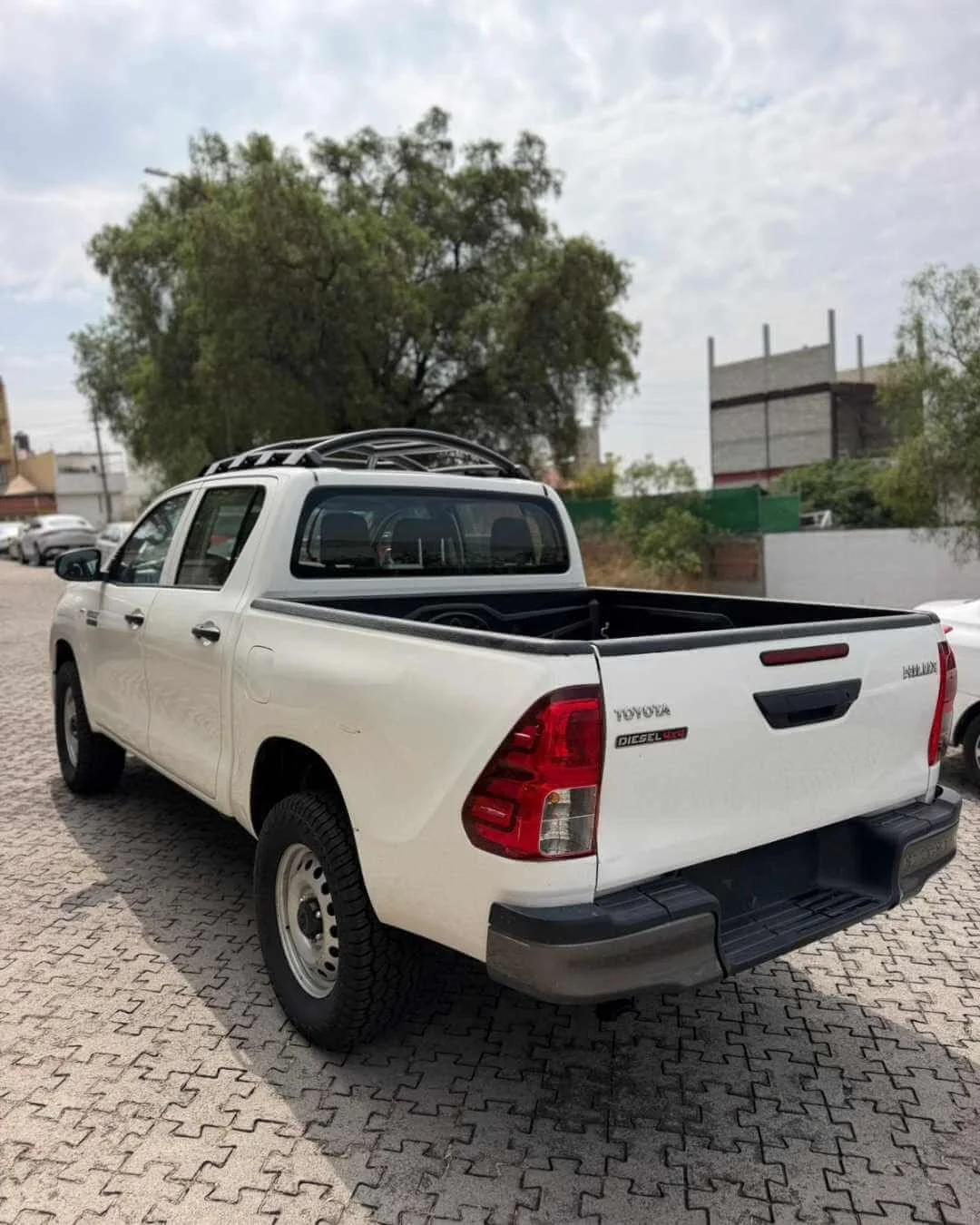Camioneta blanca estacionada en un lote con pavimento de ladrillos, árboles y construcciones en el fondo.