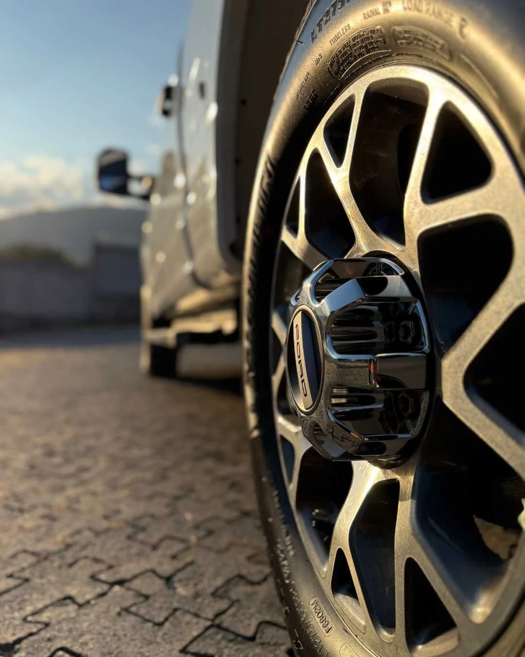 Primer plano de la rueda de un coche de carrera, con un neumático Porsche, en un estacionamiento con pavimento de bloques de piedra y un fondo borroso con un automóvil y el cielo