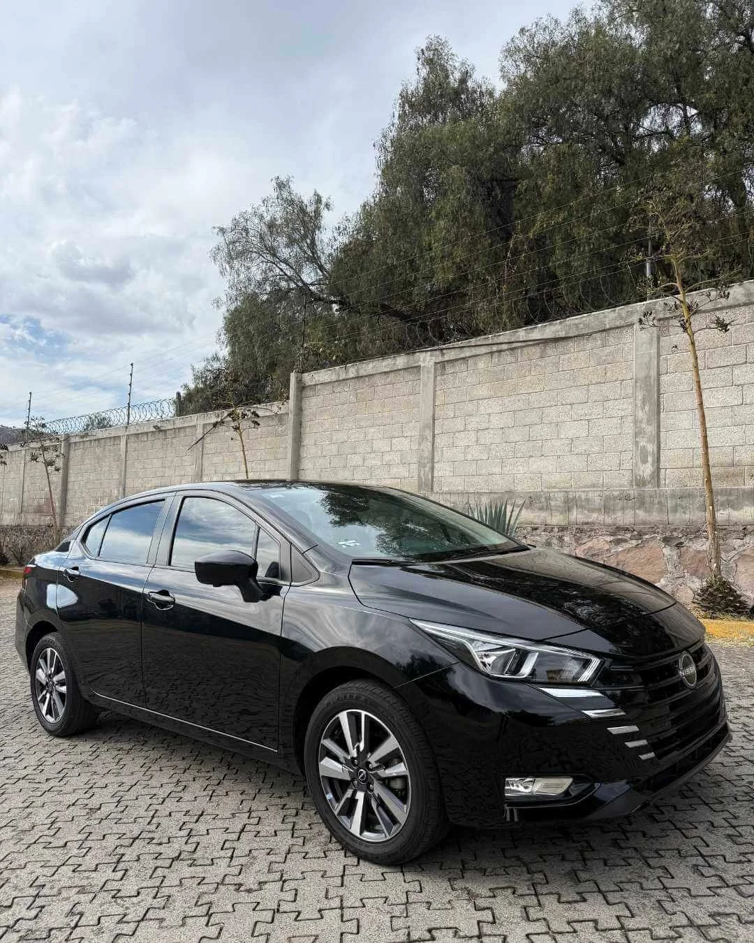 Auto negro estacionado en una superficie de bloques de piedra, con un muro de cemento y árboles de fondo