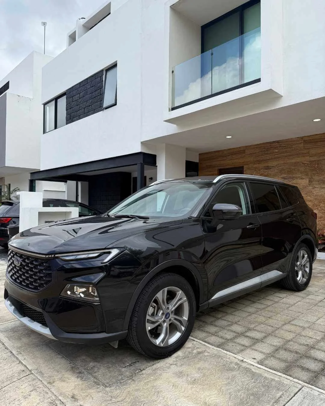 Autos negro y gris parqueados frente a una casa moderna de dos pisos con balcones de vidrio y fachada blanca.