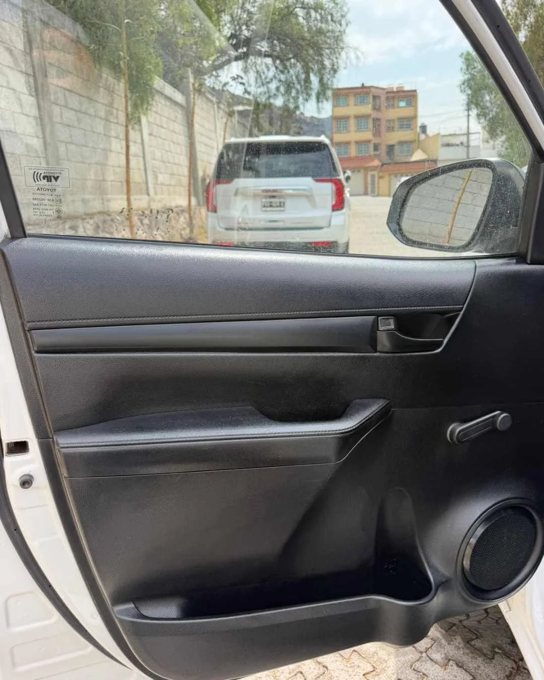 Vista del interior de una ventana del coche, mostrando la puerta con un panel negro, un espejo retrovisor y un manubrio de ventana, con un vehículo y edificios en el fondo.