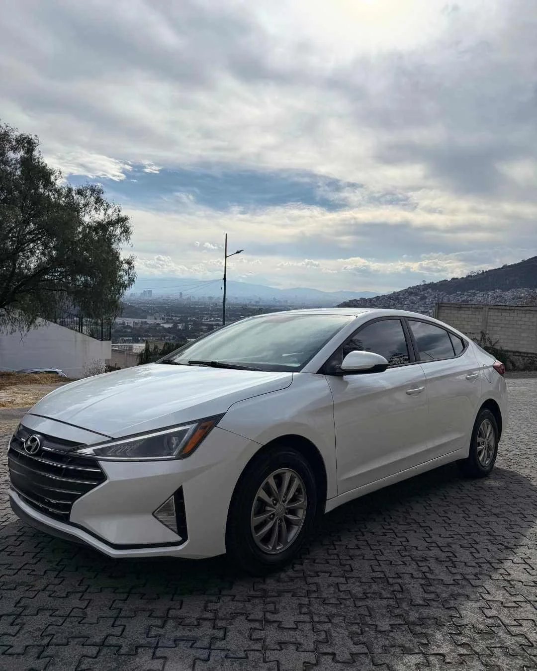 Auto sedán blanco estacionado sobre pavimento de piedra con vista a colinas y ciudad en el fondo bajo cielo nublado.