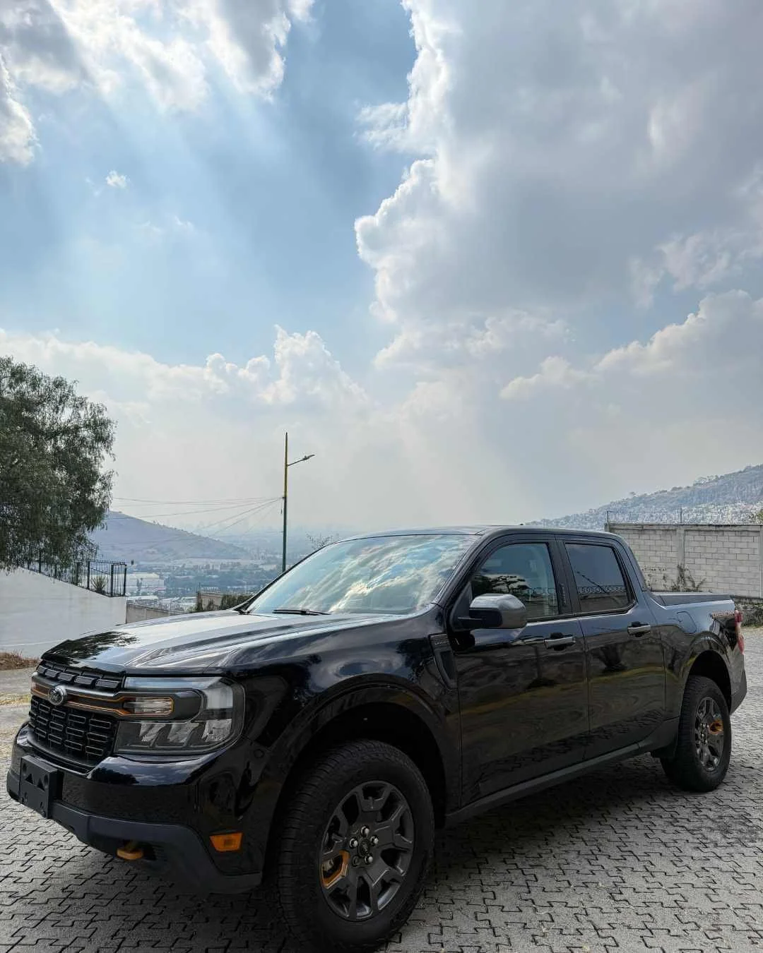 Camioneta negra estacionada sobre una calle de adoquines, con cielo nublado y colinas en el fondo.