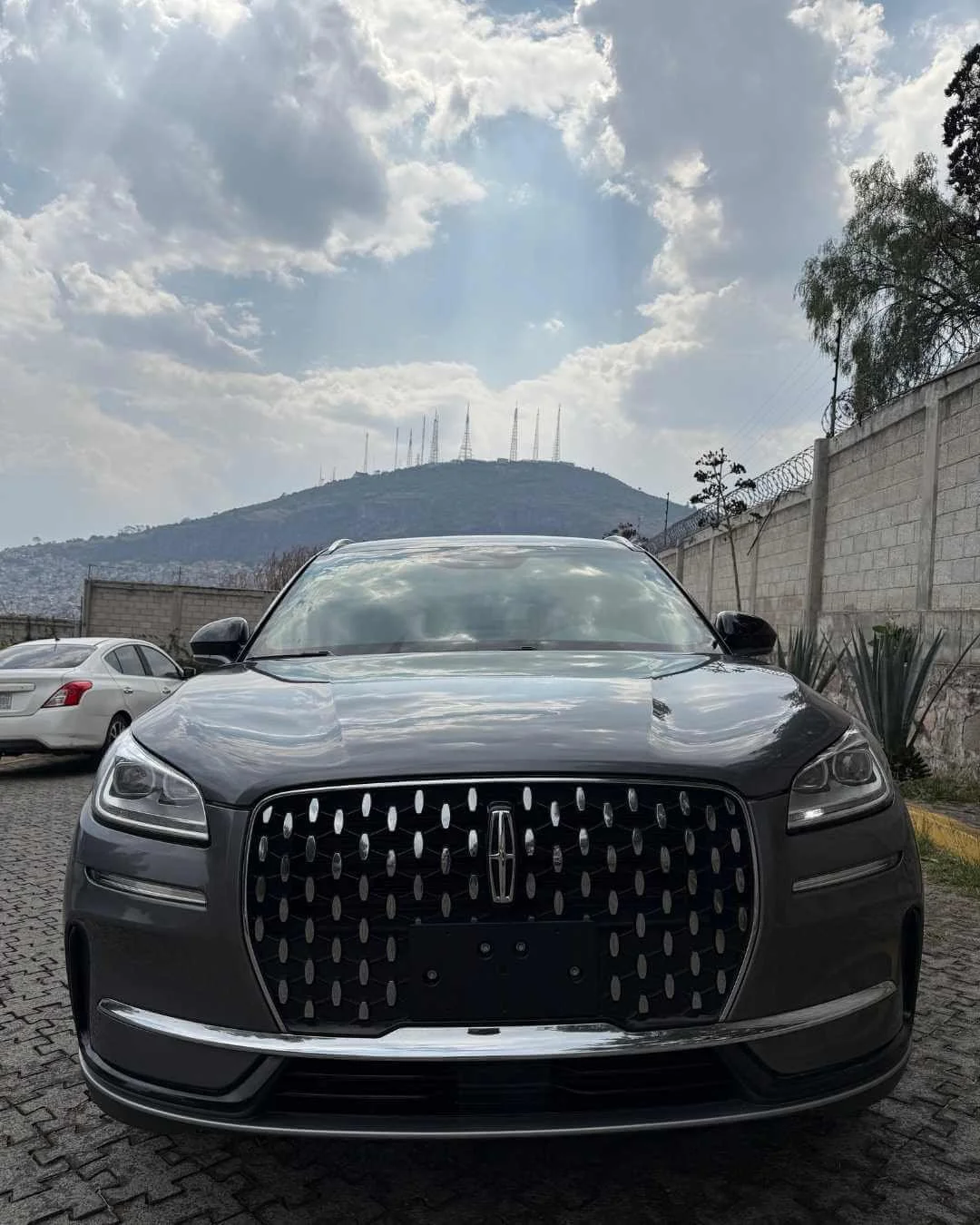 Coche negro frente, con colinas y antenas en el fondo, en un espacio abierto con otros autos y muro de bloques.